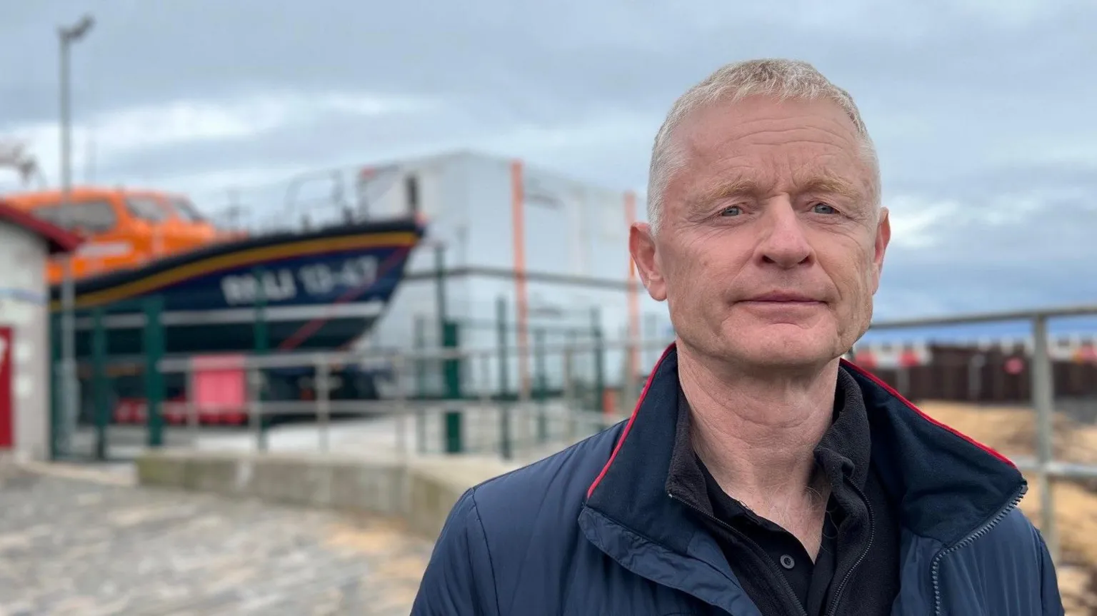 A man with short blonde hair and a blue jacket stands in front of a lifeboat