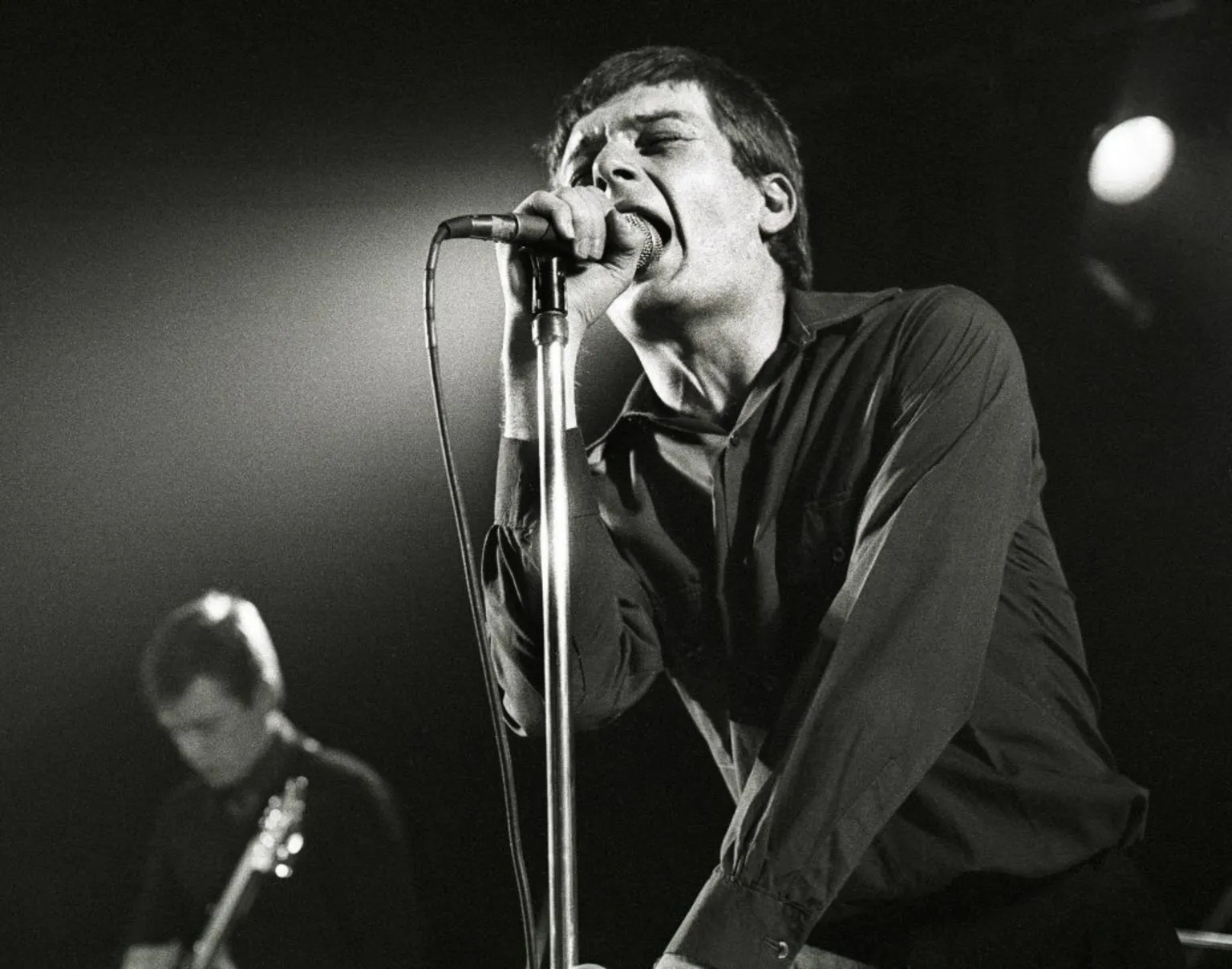 Ian Curtis of Joy Division, pictured in 1980, singing into the microphone on stage with his eyes closed.