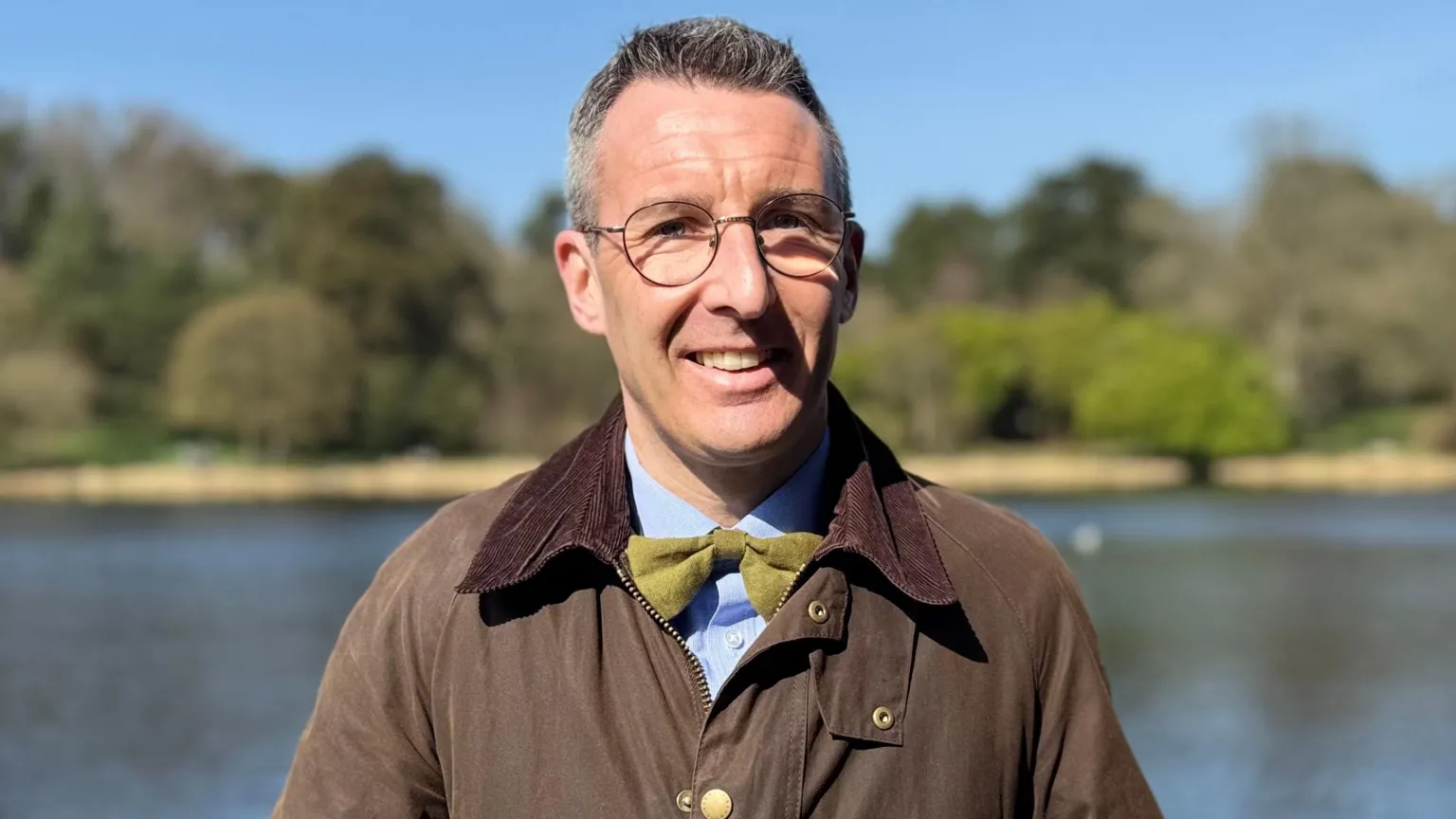 A man looking at the camera. He is wearing glasses and a brown coat, blue shirt and green bow tie