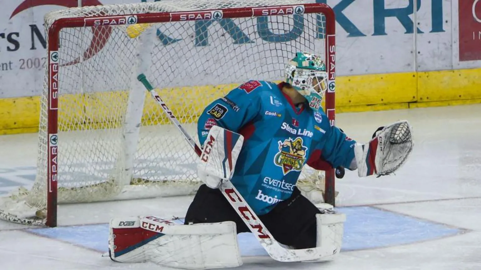 Getty A Belfast Giants goaltender makes a save with his glove. He is wearing a blue and red uniform and in full hockey mask and gear. The goaltender is on his knees in front of goal on the ice. 