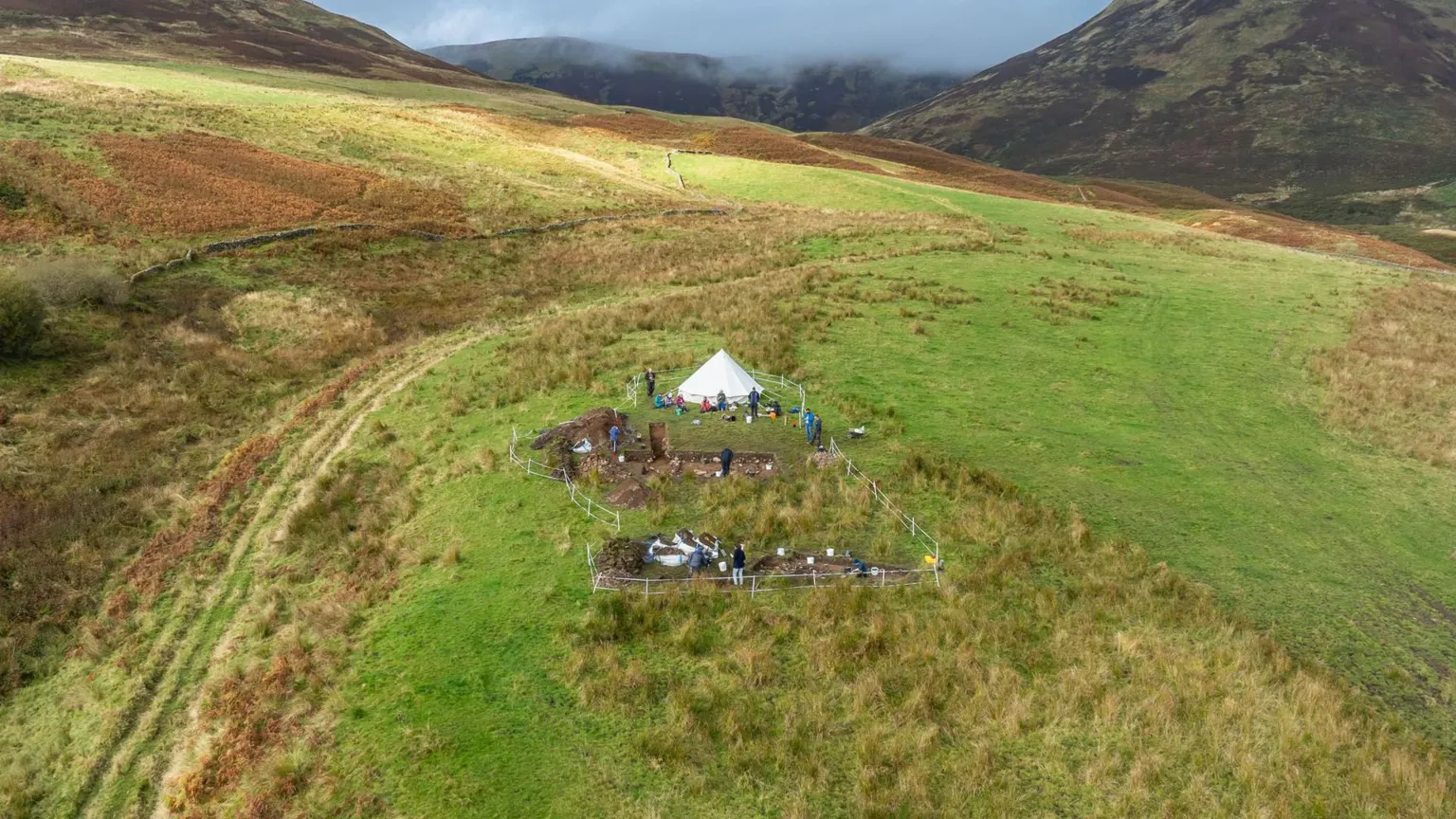 Airborne Lense/Destination Tweed An aerial image of Adie's Brae scooped settlement, showing a white teepee-style tent with two excavation pits surrounded by white tape, sitting in a hilly landscape with volunteers standing around the pits