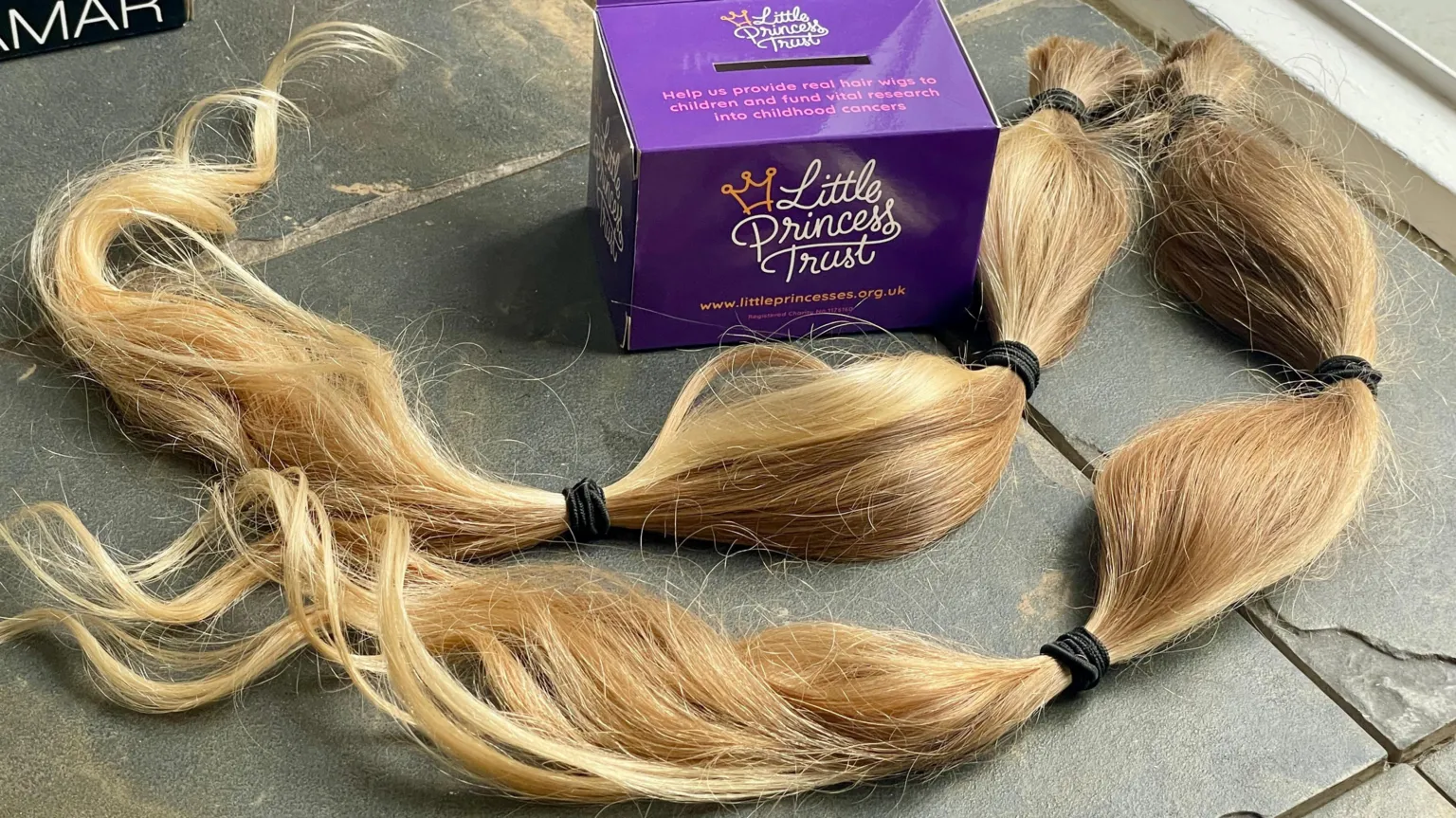 Stephen Huntley/BBC Two ponytails of blond hair, sealed with three black hair bands on each, laid out beside a purple cardboard donations box for Little Princess Trust