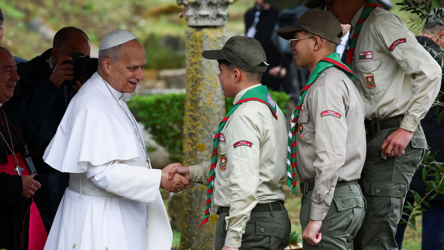  Pope standing on the left, shaking hands with a male scout. There are two others next to him. They are standing outdoors. Behind the pope is a man holding a camera up to his face.