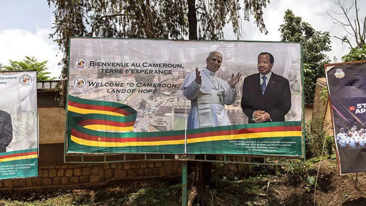 AFP via Billboard in the centre, with slogan welcome to Cameroon, land of hope on the left and a ribbon acorss the lover half of the billboard in - green, red and yellow horizontal stripes. On the centre right is the Pope with his hands up, and next to him is the president.