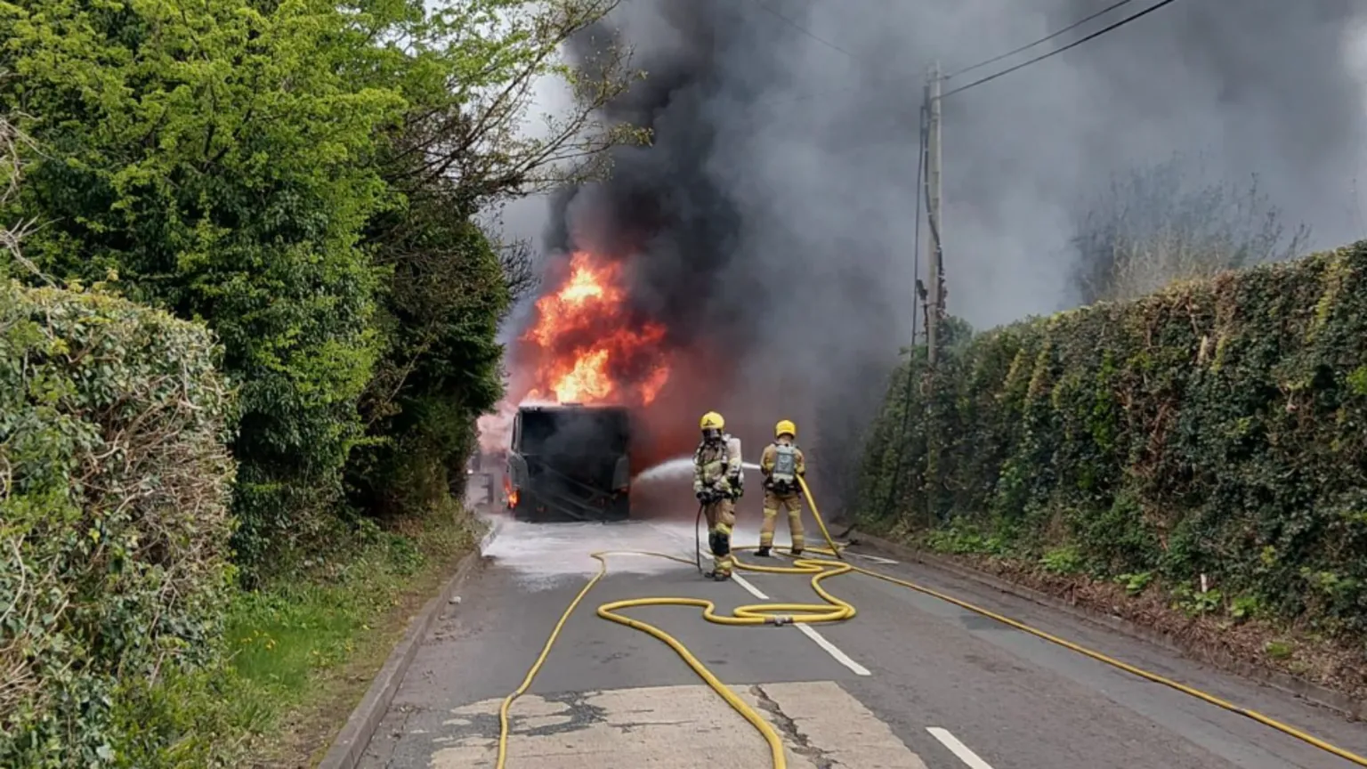 Shropshire Fire and Rescue Service Two firefighters in light brown clothing and yellow helmets directing long yellow hoses at a burning object on a road with bushes on either side