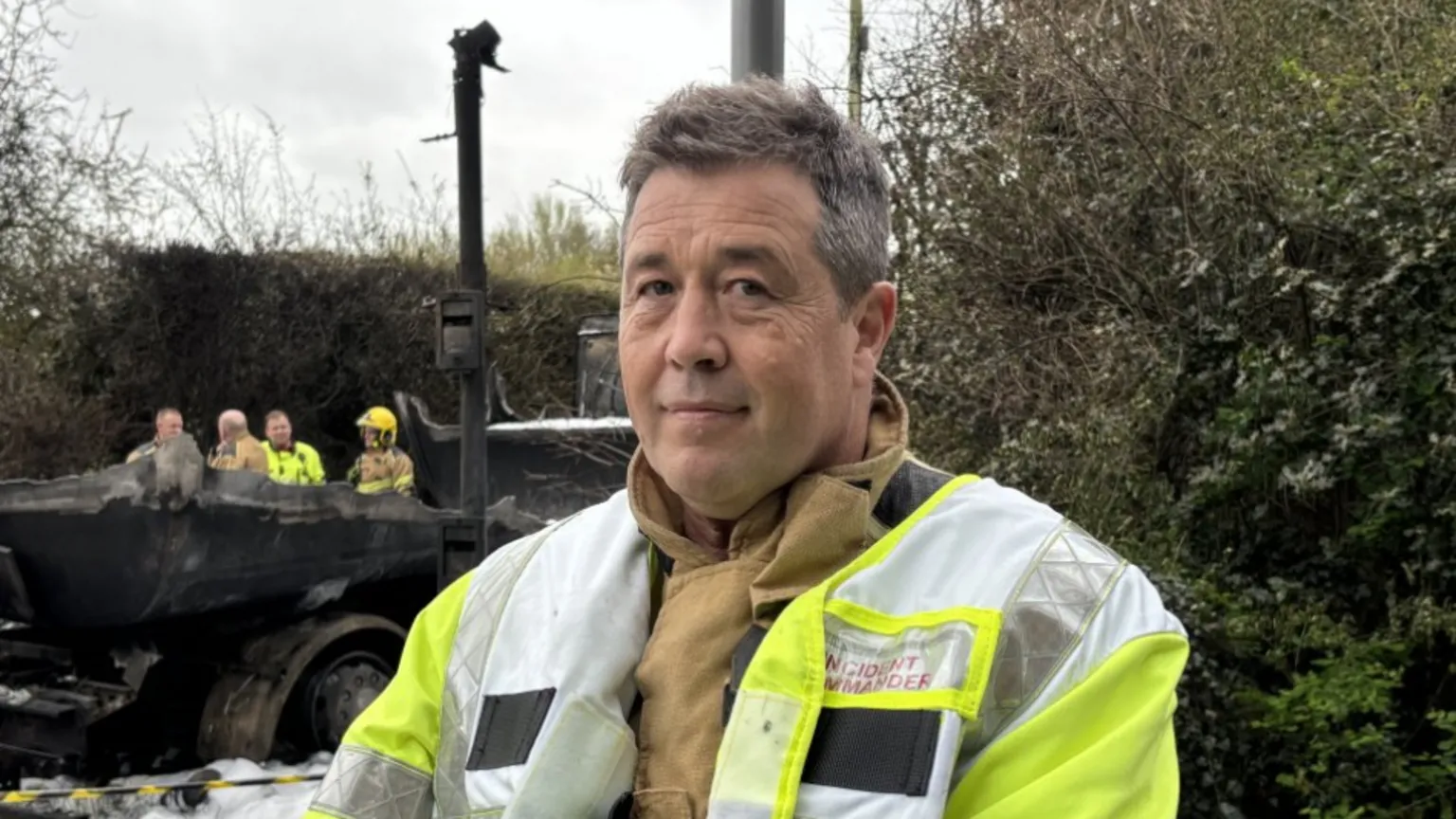 A man with short grey-black hair and a yellow reflective jacket standing in front of bushes with a black, burnt-out vehicle behind him