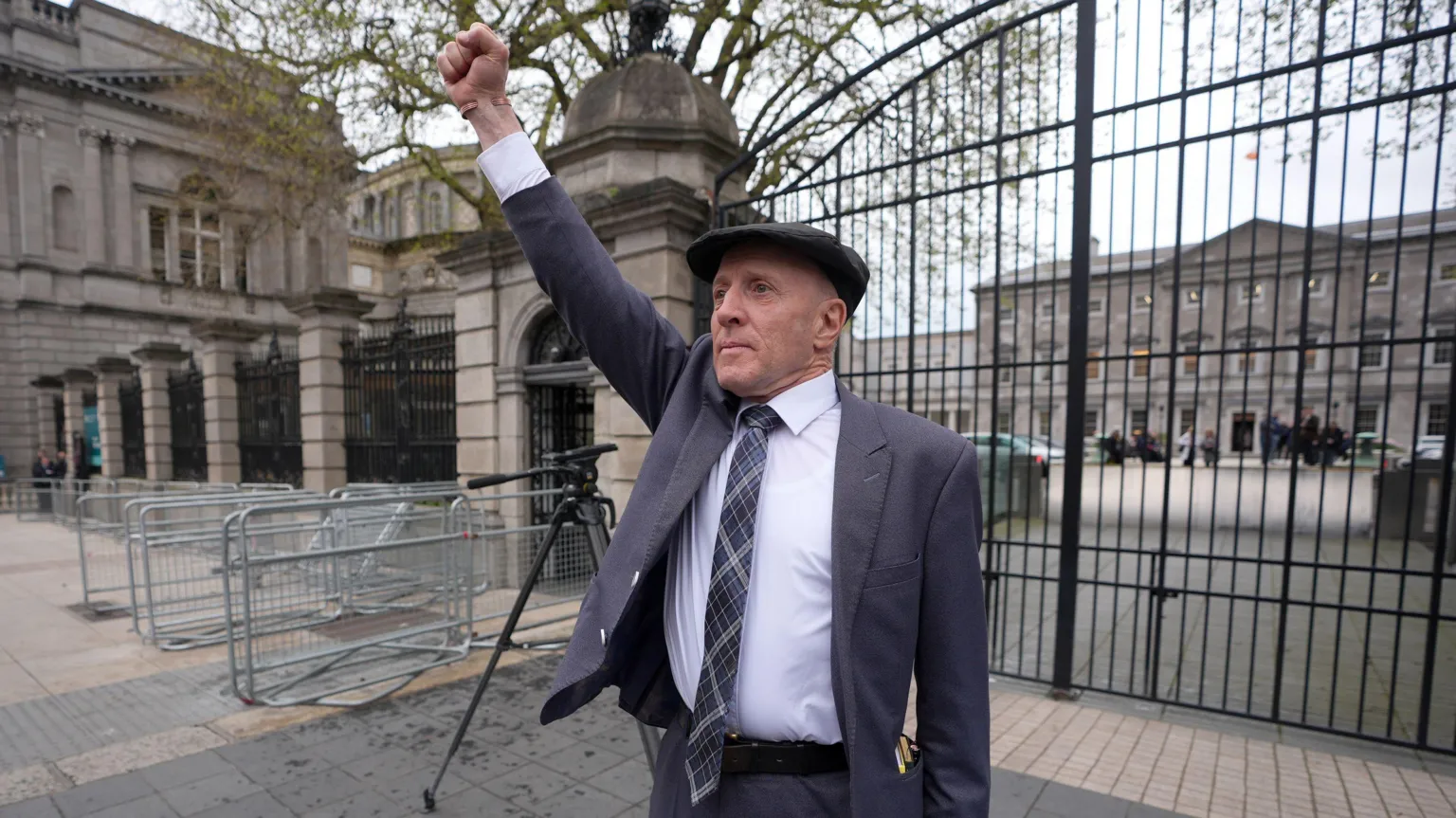  Michael Healy-Rae, a man with shaved head and a flat peaked cap, stands outside the tall iron gates of Dublin's Leinster House, with one fist raised above his head. He is wearing a grey/blue suit, a light shirt and a navy tartan tie. 