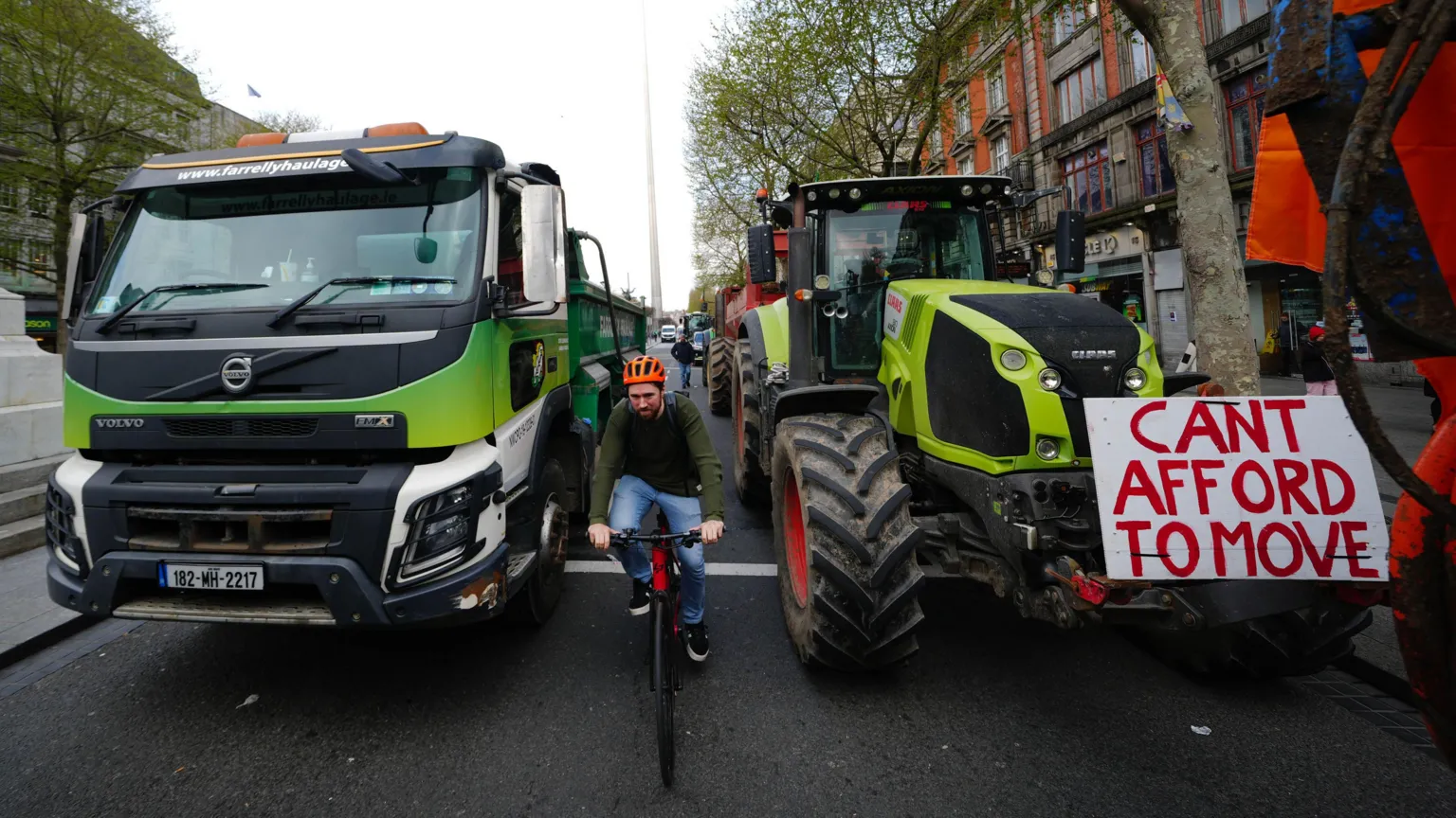 Brian Lawless/PA Wire A cyclist wearing an orange helmet is between two tractors on a main city street. A handmade sign on the tractor says: 