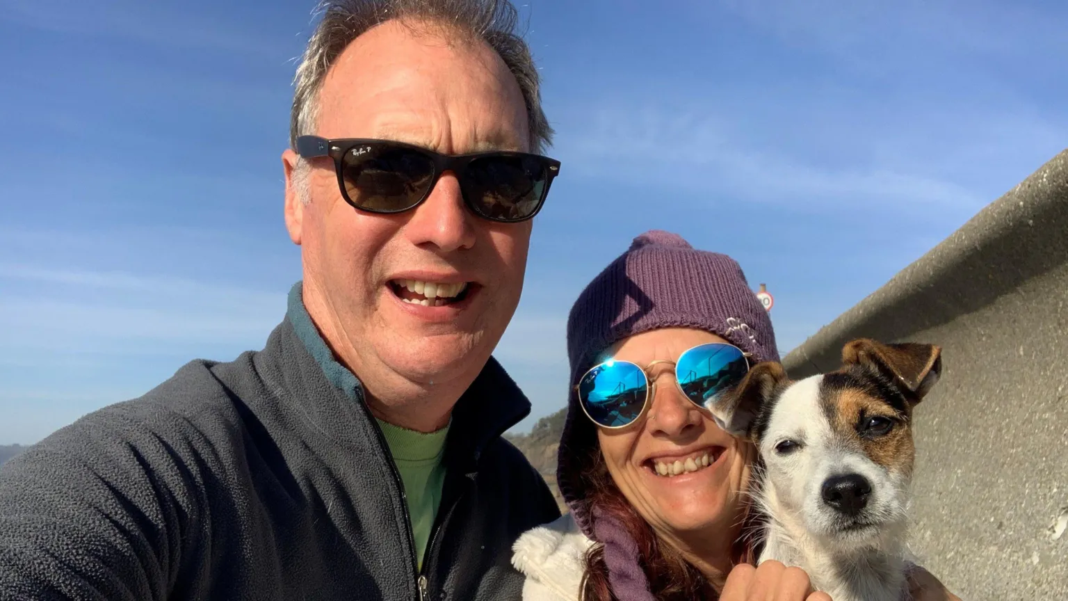 Martin Johnson A grey-haired man with black sunglasses, wearing a blue fleece and holding his arm out to take a photo in selfie-mode. To his right, a brunette woman, wearing a purple hat and blue sunglasses. She holds her Jack-Russell dog in both hands. 