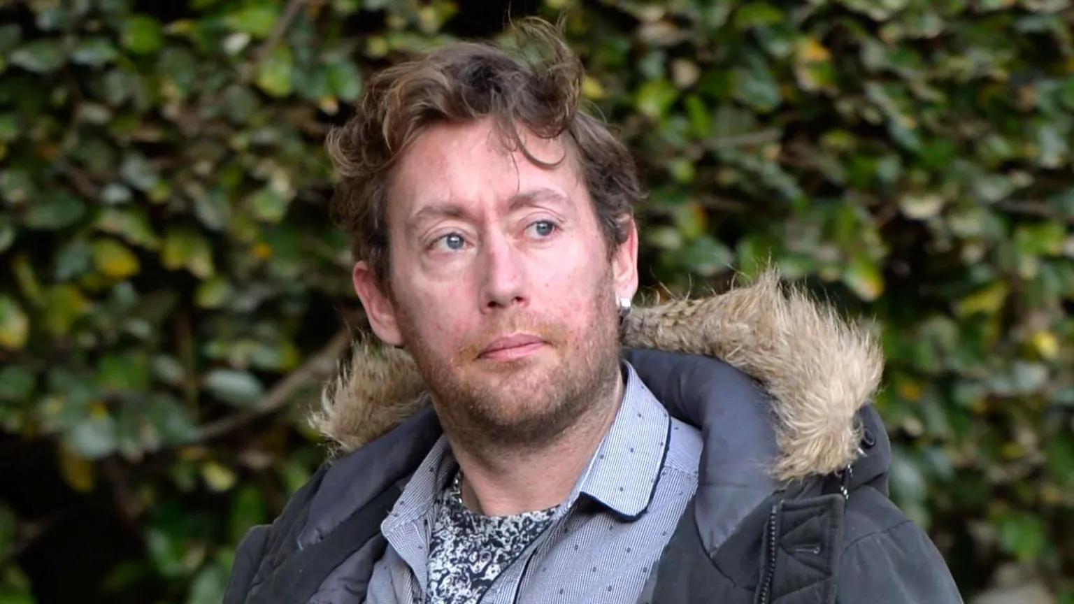 Spindrift Lee Milne is in his late 30s with wavy brown hair. He is wearing a blue parka jacket and a blue shirt. There are bushes in the background and he is looking to the right of the camera.