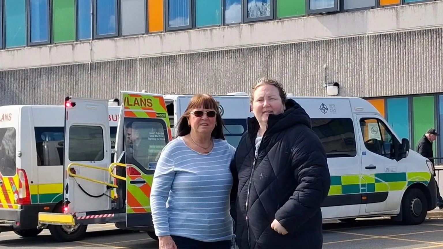 Delia Lodge Delia is stood on the left and Rebecca is stood on the right. They are in front of ambulances at Ysbyty Glan Clwyd Hospital's A&E department.