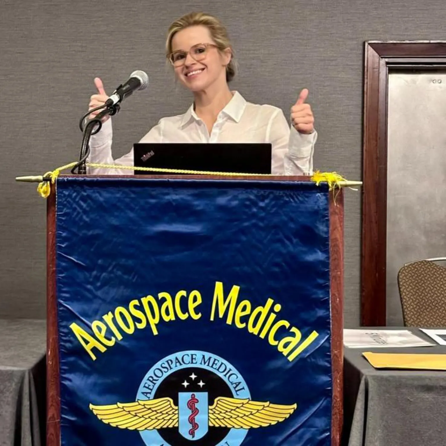 Christina Mackaill Christina Mackaill standing at a podium behind a flag reading AEROSPACE MEDICAL ASSOCIATION. She is holding her thumbs up wearing glasses and a white blouse.