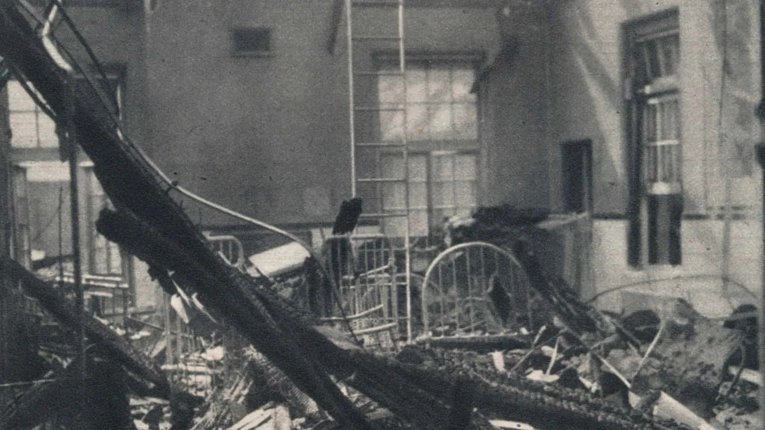  A black and white archive photo showing rubble lying in a hospital ward. Beams from the roof are lying across thew floor. Beds are damaged. Debris is covering the floor. 