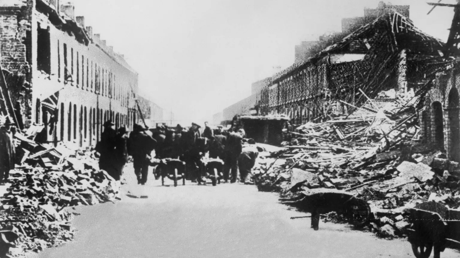  Rubble is on the sides of a street. Houses are damaged and brick and timber are exposed. People are trying to clear some of the debris. 