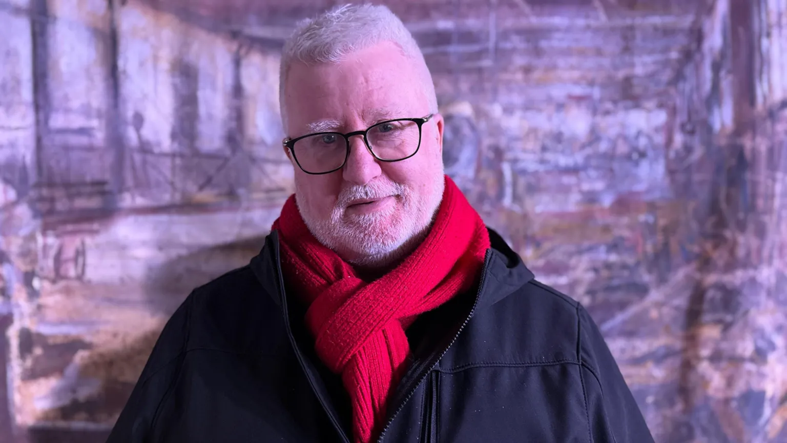 Trevor is looking into the camera and smiling. He's wearing a red scarf and a dark navy jacket. He wears glasses. A backdrop is behind him showing a factory scene. 