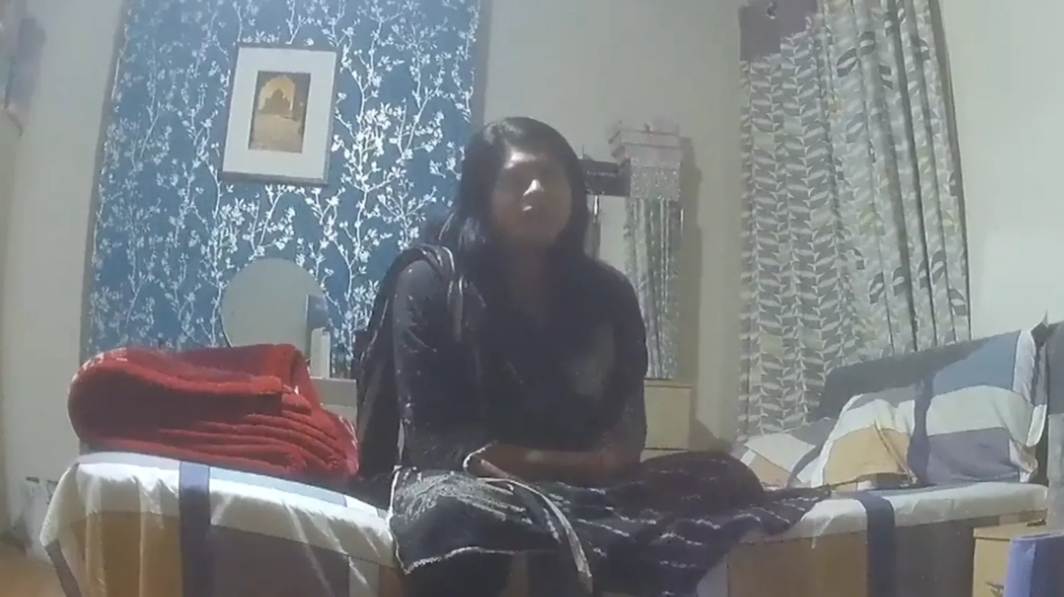 Undercover footage of an Asian woman sitting on a bed. The chimney breast behind her has been turned into a feature wall with bright blue and silver wallpaper, while the bed cover is a large check pattern in yellow, blue and grey.