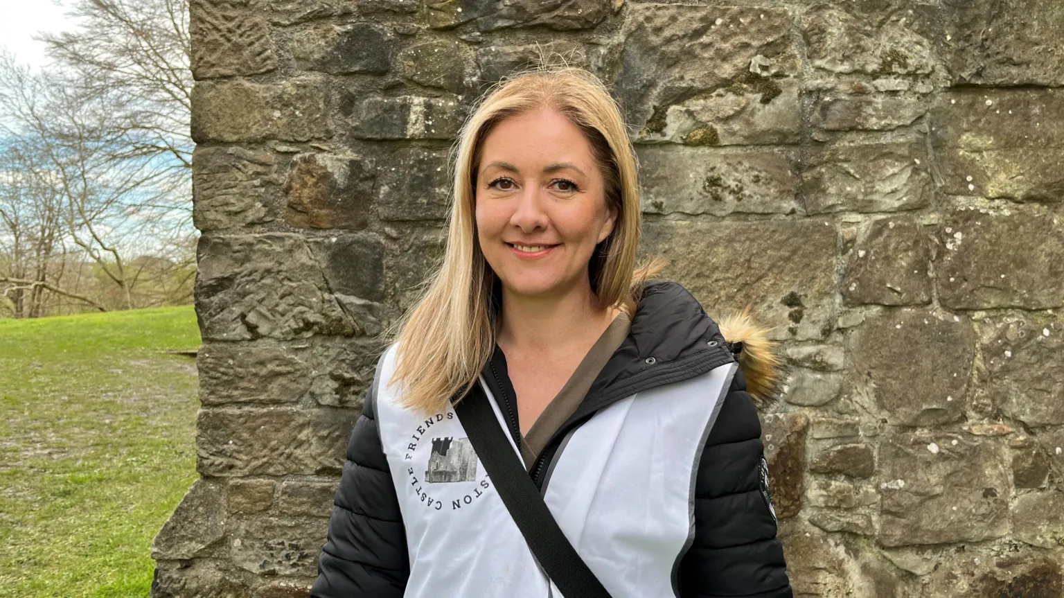 Trisha Robertson - a woman with long blonde hair and wearing a white bib saying Friends of Crookston Castle, standing in front of a castle