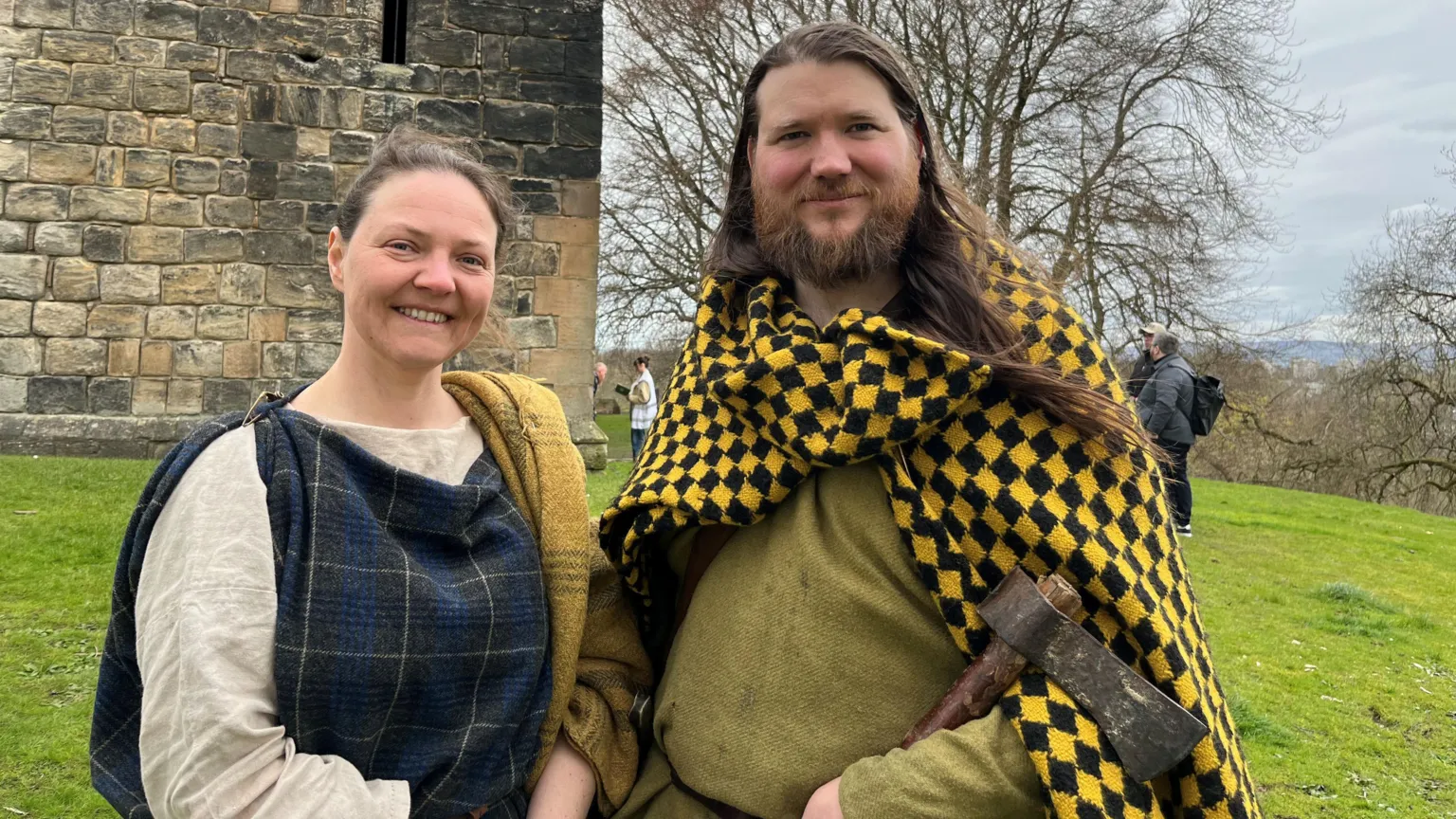 Caroline Nicolay and Thomas Timbrell - a pair of living history reenactors dressed in Iron Age clothes - standing in front of a castle. Thomas has an iron age axe in one hand. 