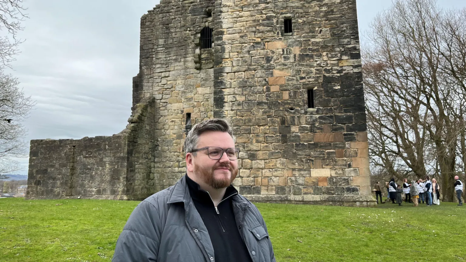 David McDonald - a man standing in front of a castle, with glasses, short dark/greying hair and a grey jacket and black top.