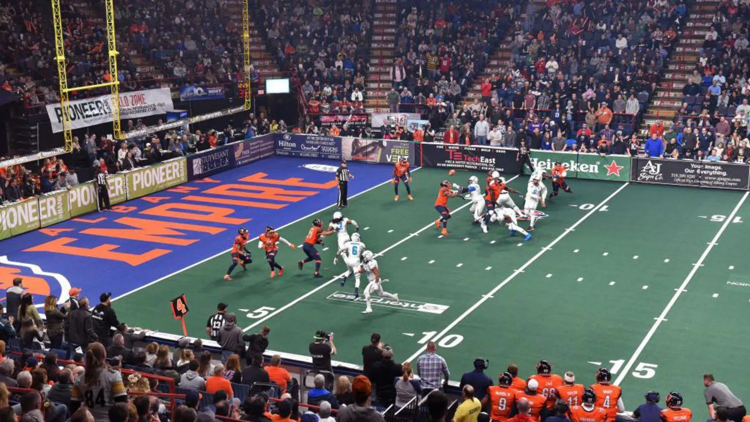  An indoor American football game, with two teams competing - one in mostly white and the other in orange and black.