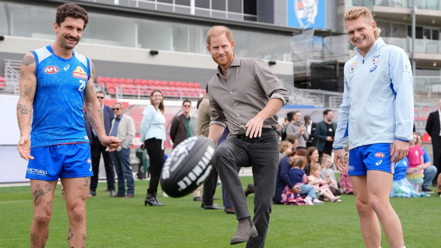 Prince Harry stands next to two AFL players. He kicks a black ball with white text on it into the foreground.