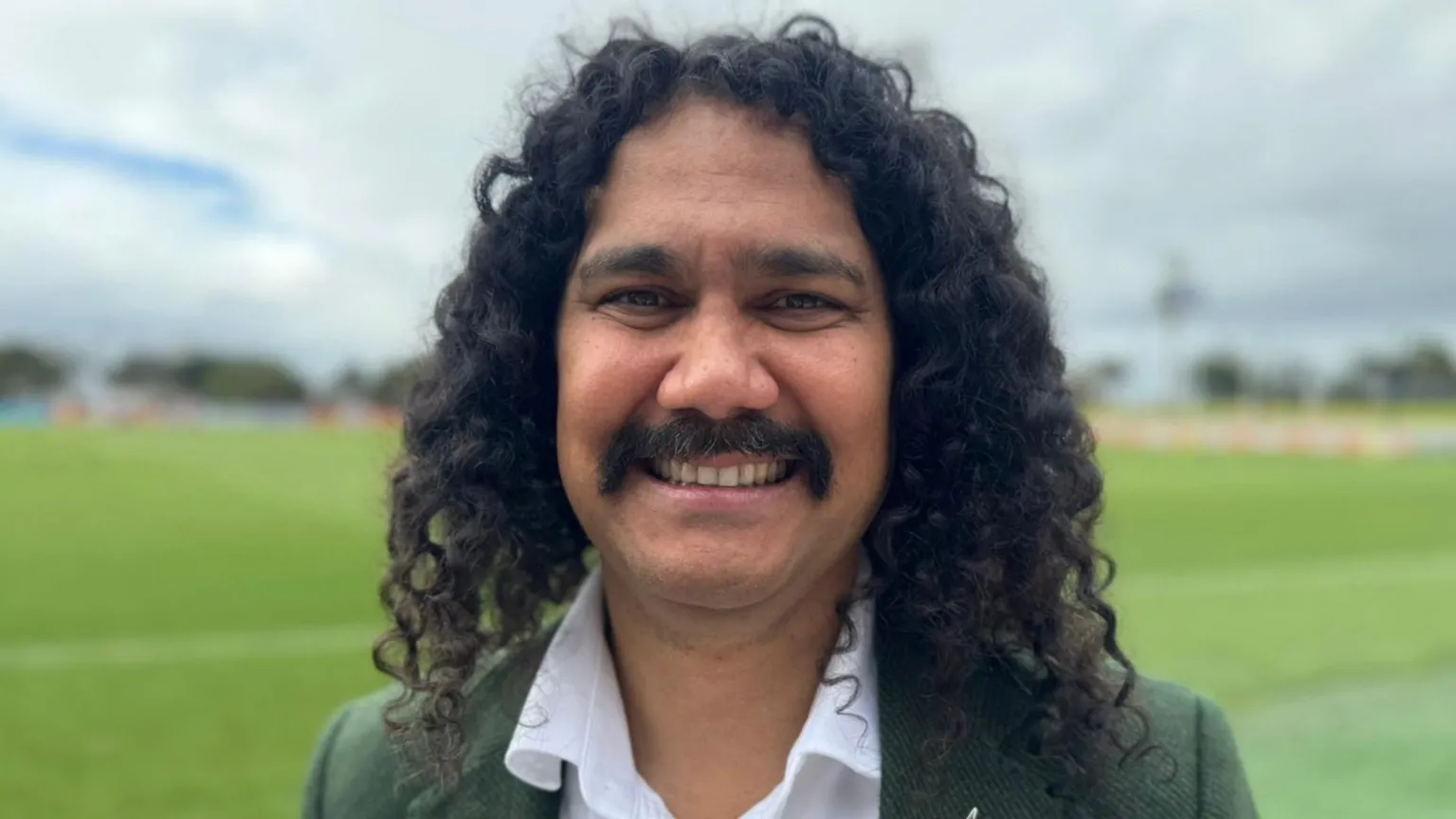 Nathan Appo, a man with dark curly hair and a moustache grins. He wears a green jacket and white shirt and stands in front of a sports field.