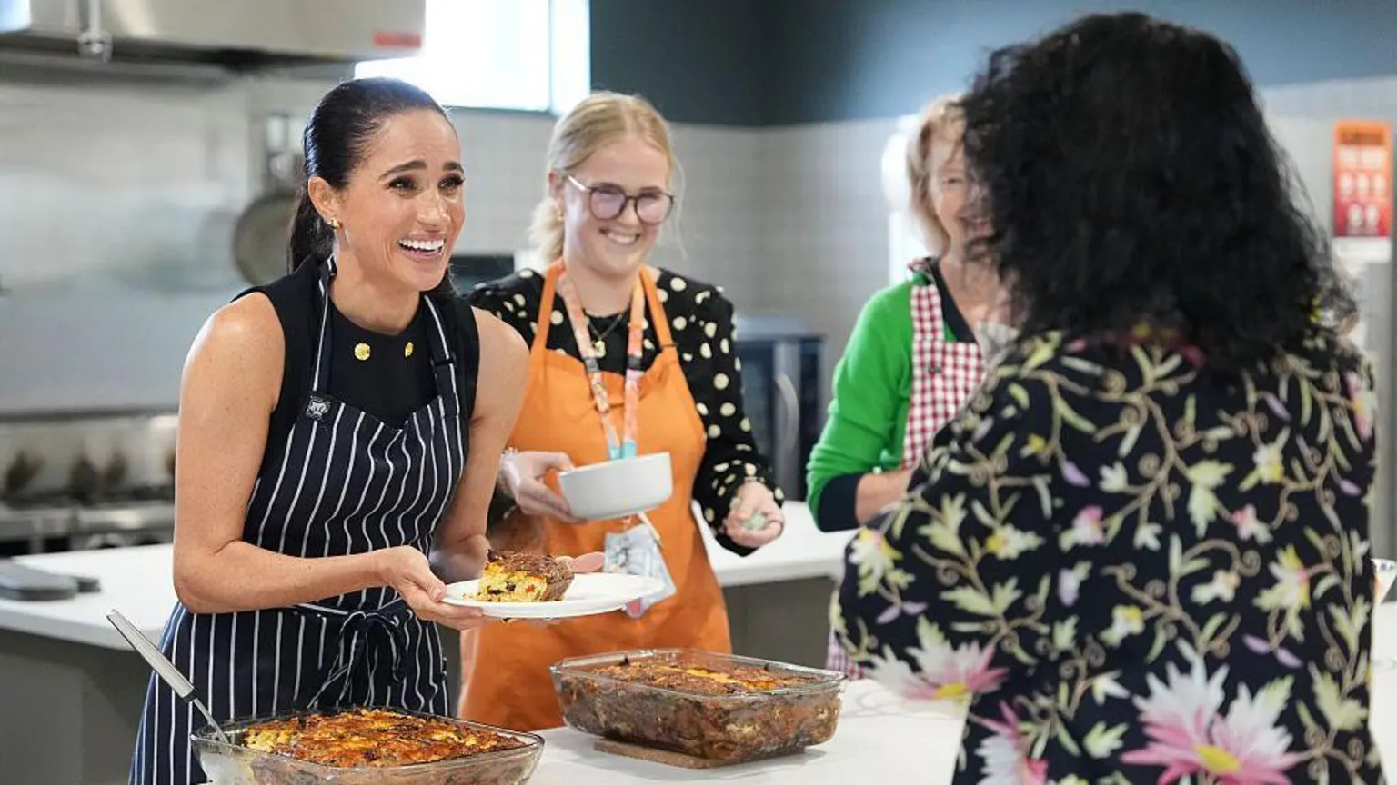 Duchess of Sussex smiles as she presents a plate of food to another woman in a floral-patterned shirt. Two other woman grin next to her.