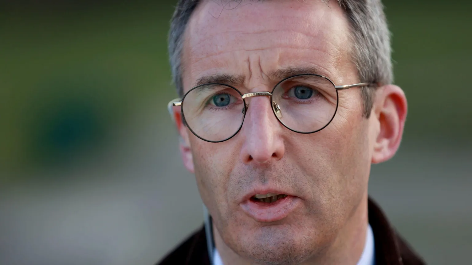  Andrew Muir, a man with short grey hair and blue eyes, speaks to the media. He is wearing large, round metal-framed glasses and a brown jacket over a pale shirt. The background of the close-up photo is blurred.