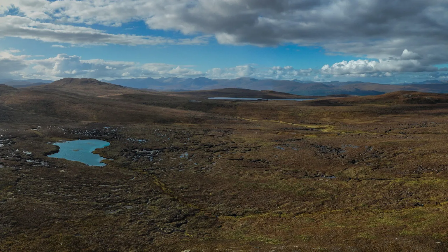Tracks Ecology Limited A wide open stretch of peat bog and moorland. A large pool can be seen on the left hand side of the picture. There are no trees to be seen. The sky is a dramatic with blue colours and white and grey clouds.