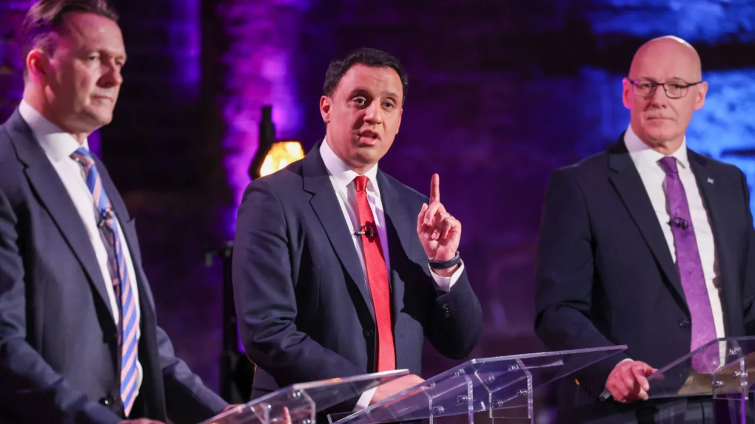 Three men in suits stand at lecterns during a debate; the central speaker in a red tie raises a finger while speaking, flanked by two others, against a purple-lit brick backdrop.