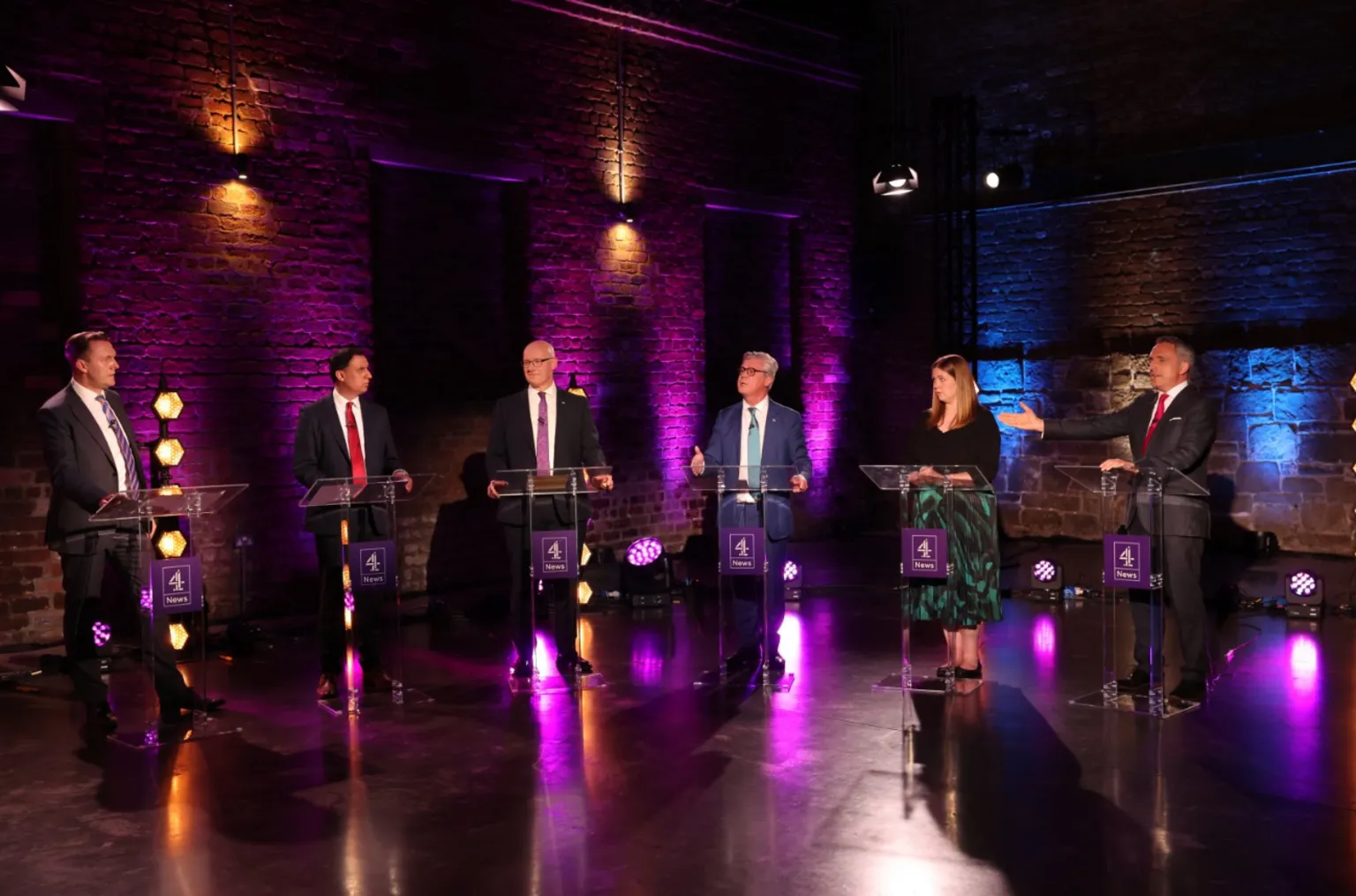  Six people stand at lecterns during a televised debate in a studio with purple and blue lighting against exposed brick walls; one speaker gestures toward another while the others look on.