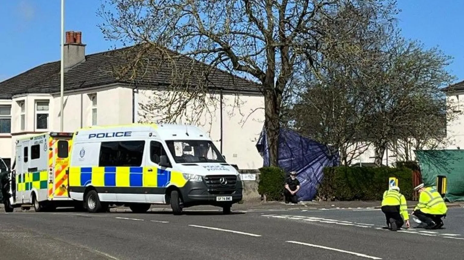 Police and ambulance outside a white property on the junction of two roads. Police officers are checking the road.