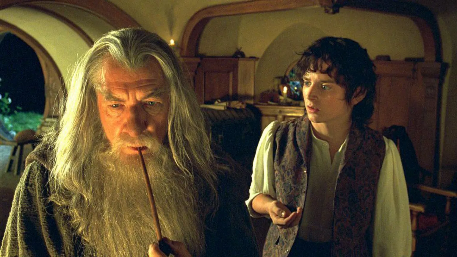 New Line via Ian McKellen on the left is wearing a grey hooded robe holding a smoking pipe to his mouth. He has long grey straight hair and a long grey beard. Elijah on the right is looking at Ian. He has brown curly hair and is wearing a white shirt and open brown waistcoat.