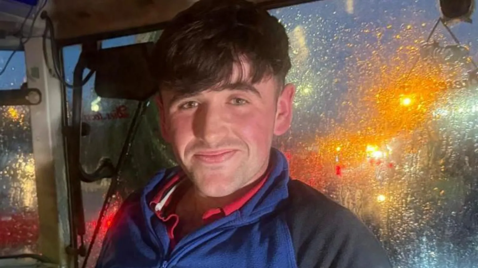 Ryan McKenna is looking at the camera. He is sitting in the cab of a tractor wearing a navy blue fleece, he has brown hair.
