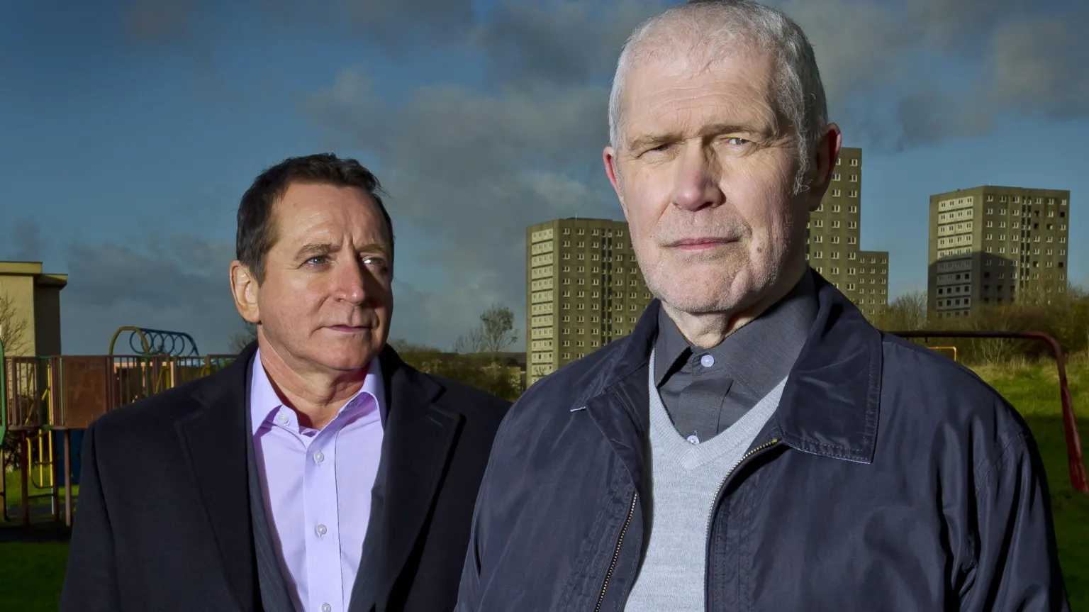 Alexander Morton on a blue jacket and jumper with a grey look serious while Frank Gallacher stares at the back of his head. There are a number of tower blocks behind them.