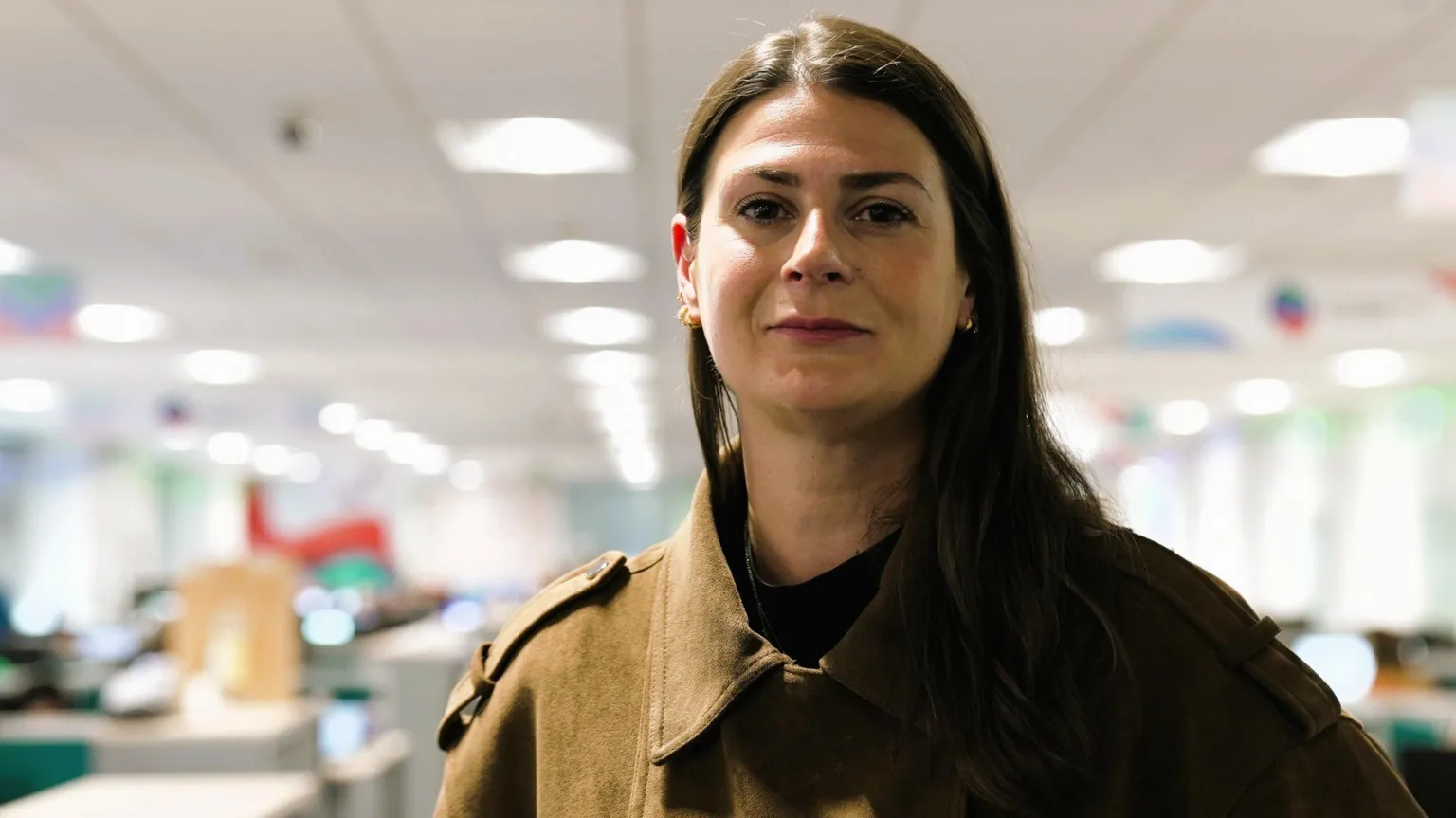 A woman stares at the camera. It is a head and shoulders shot. She has long, dark hair and has a brown jacket on. She is in focus, while the background is blurred.
