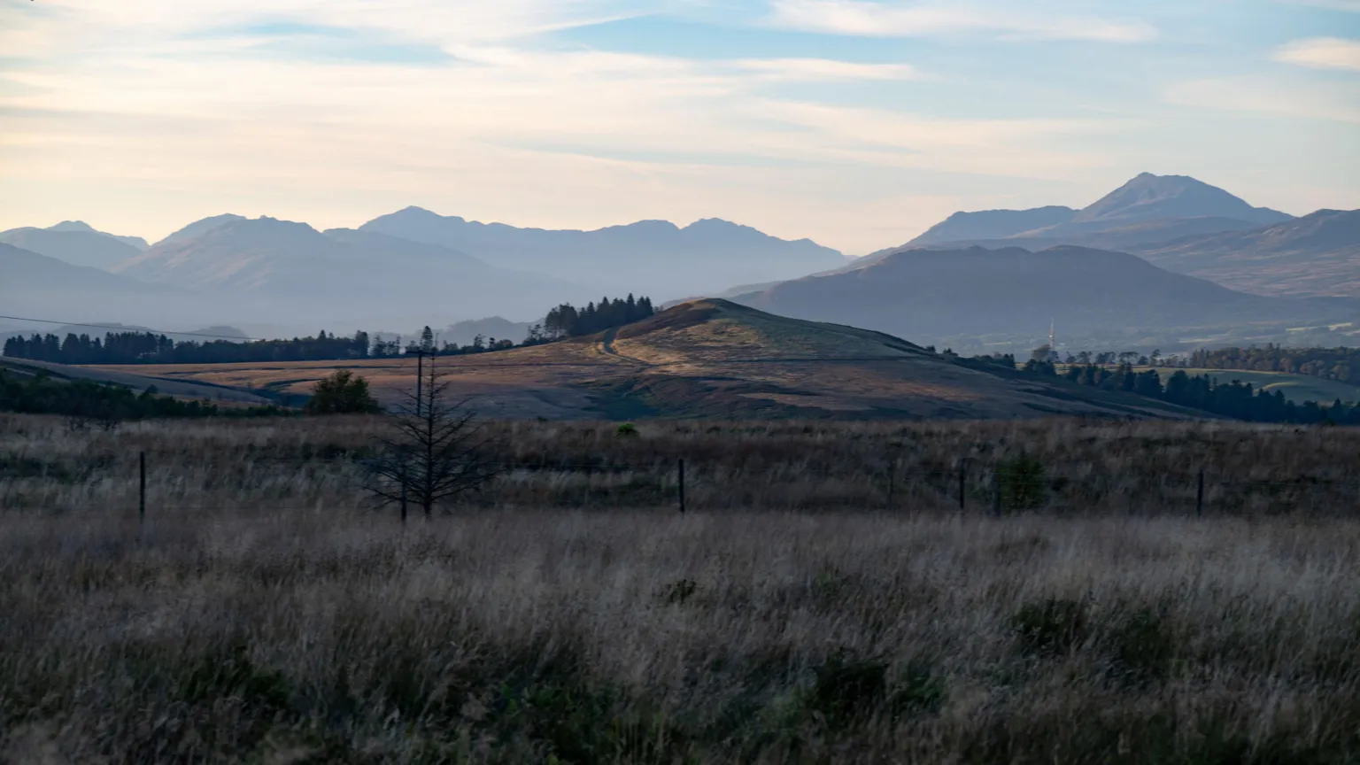 Lisa Birch A beautiful image of Scotland shows a wide, open grassland stretching across the foreground, dotted with sparse shrubs and muted earth tones. Rolling hills rise gently toward layered mountain ridges fading into the distance. Soft daylight and pale clouds create a calm, expansive atmosphere over the landscape