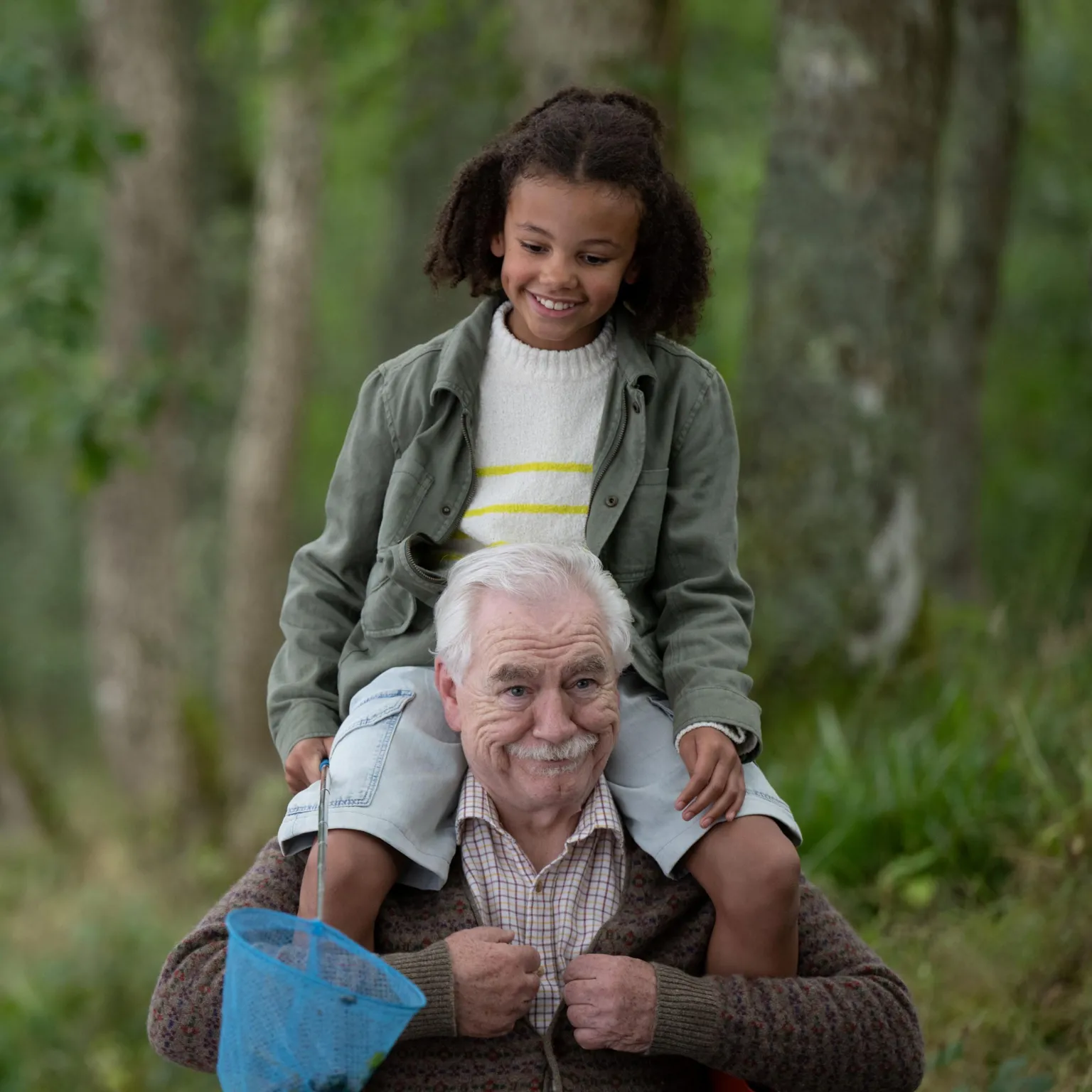Lisa Birch An image of two people outdoors in a wooded setting, surrounded by trees and soft green foliage. A young girl sits on her grandfather's shoulders, both dressed in casual, practical clothing suited to the outdoors.