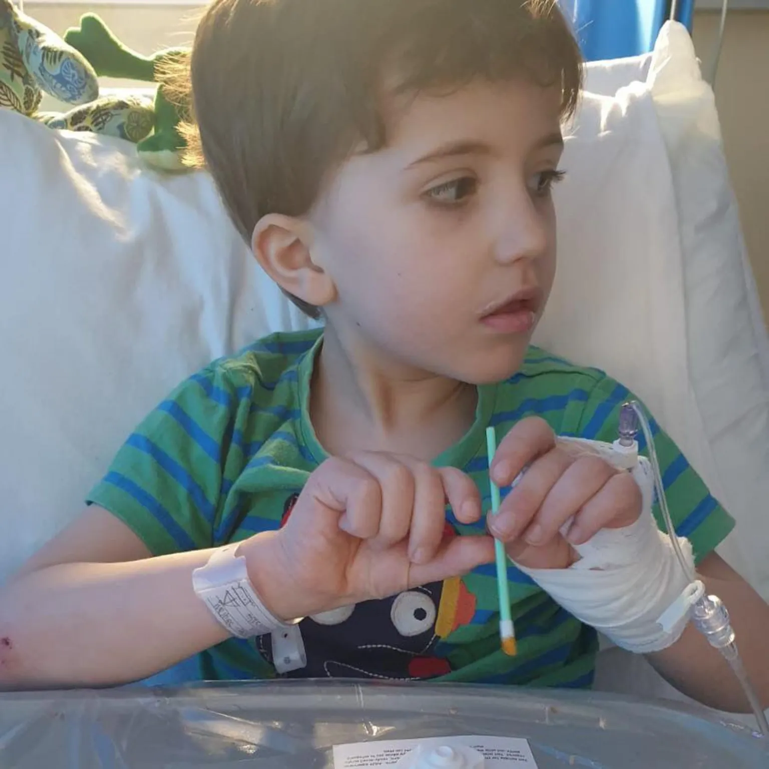 Carys Hobbs-Sargeant A young child sitting up on a bed, with a bandaged hand and a medical tube attached, holding a small swab.