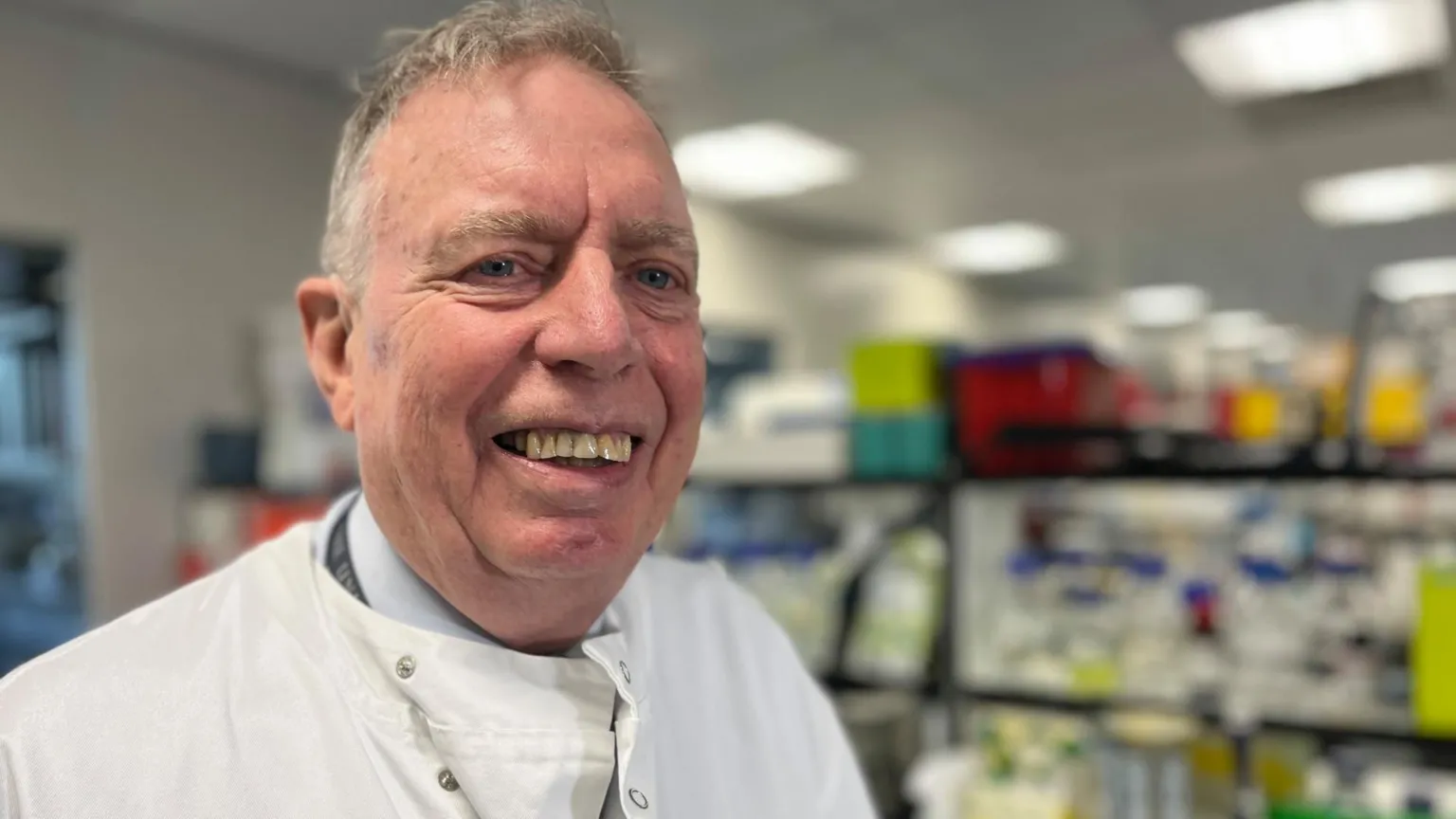 Prof Stuart Ralston - wearing a white coat, standing in a laboratory with shelves of equipment behind him