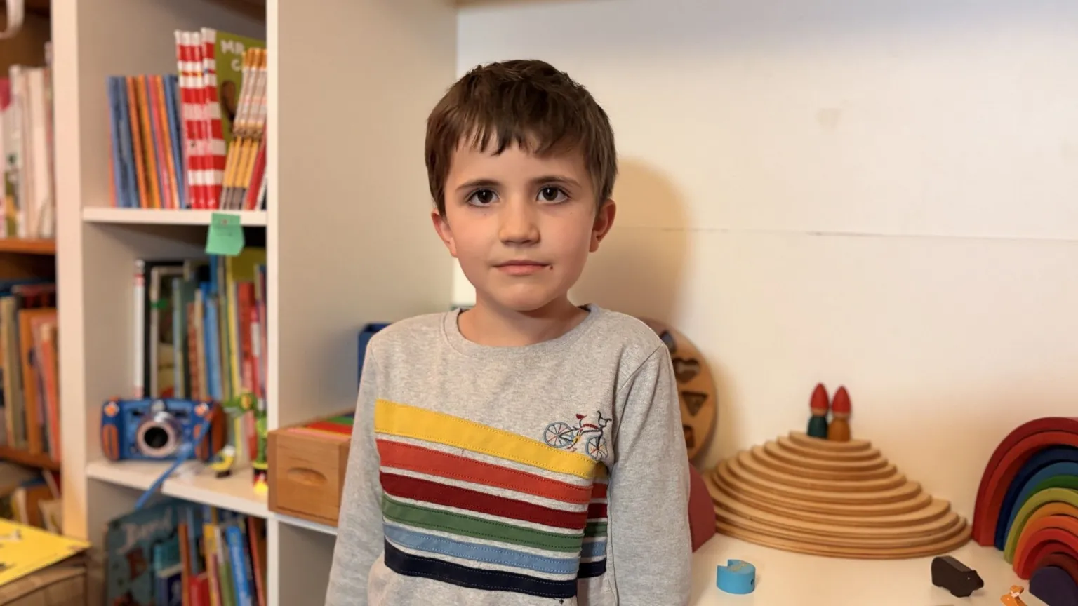 A young child standing indoors in front of shelves of books and wooden toys.