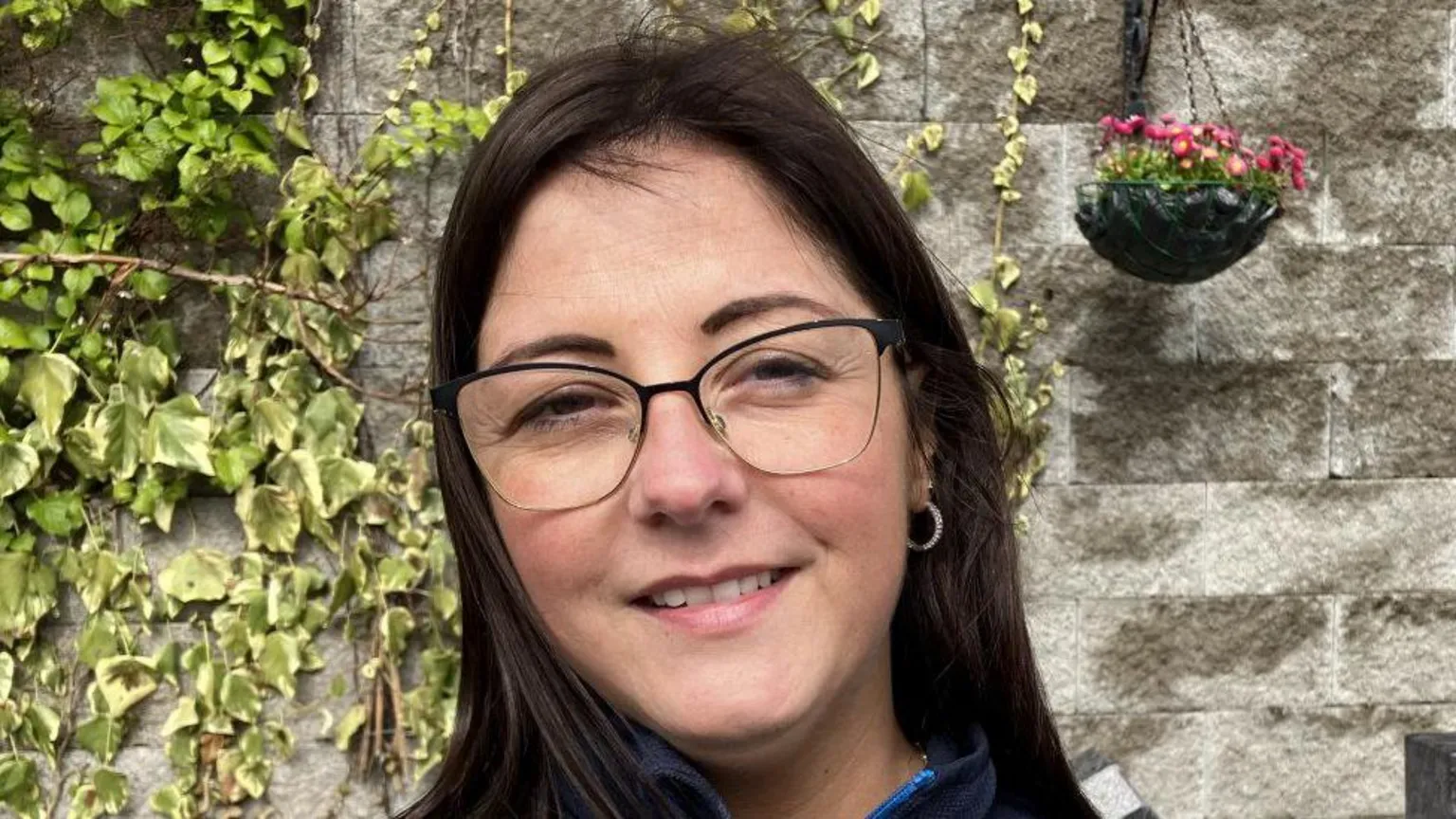 Shows a young woman with dark long hair and glasses smiling. She is standing in front of a wall, with ivy and a hanging flower pot to the right of the picture. 
