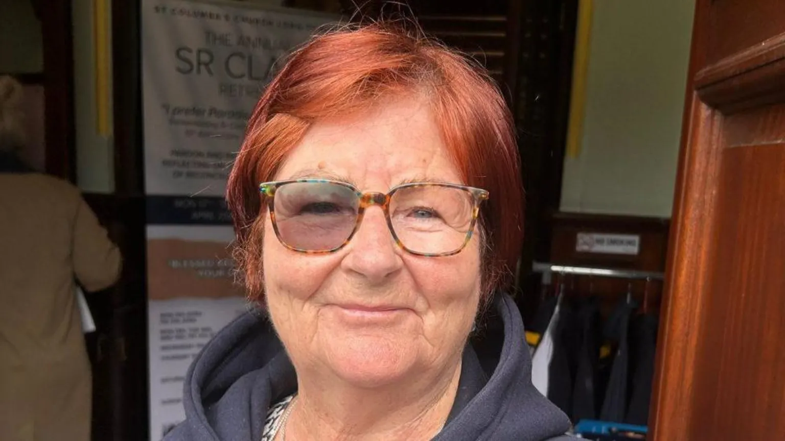 Shows a woman with red hair and glasses smiling. She is wearing a blue hoodie.