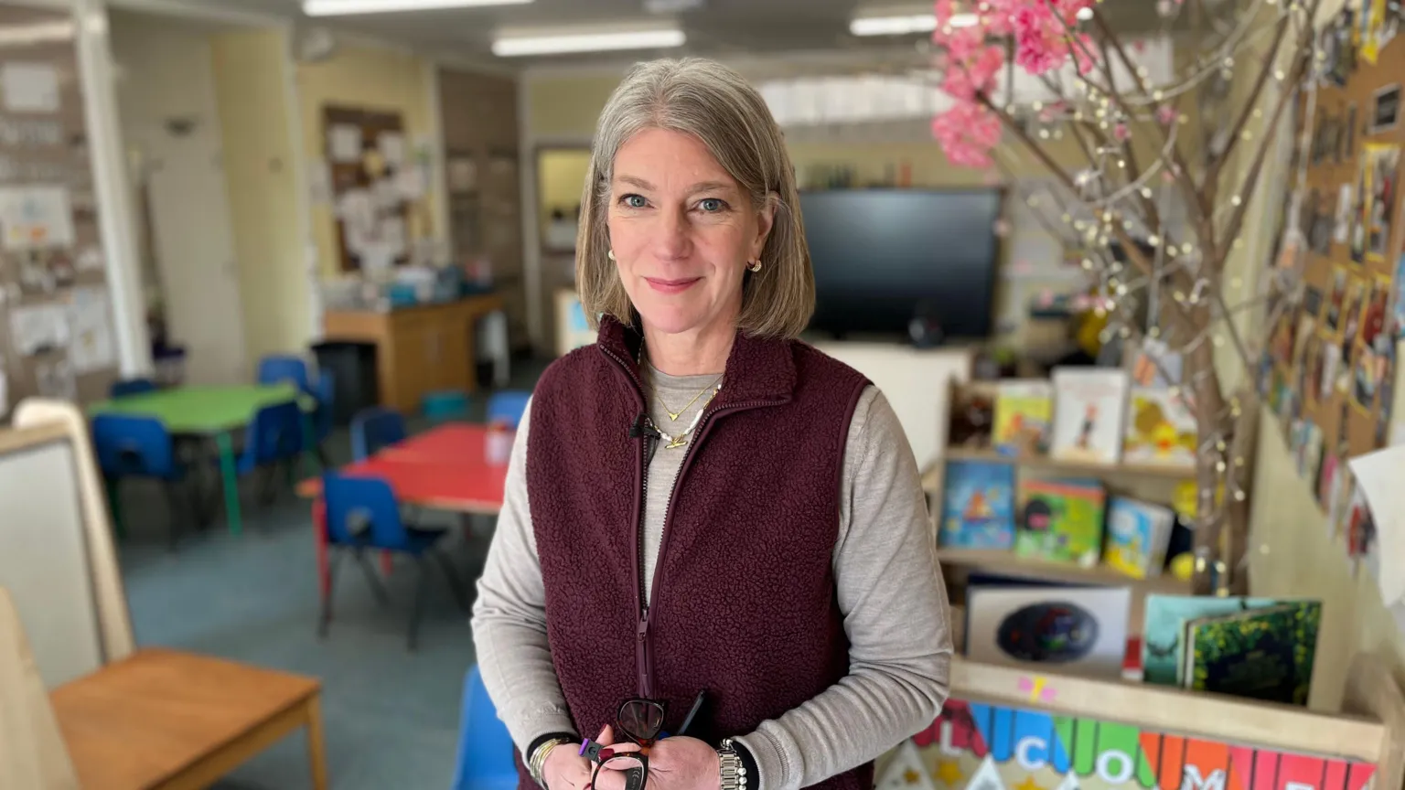 John Boon / BBC Jo Mould standing in a classroom, wearing a burgundy fleece vest over a light long-sleeved top, holding glasses. Behind are children’s chairs, tables, bookshelves with picture books, colourful displays and a decorative branch with pink blossoms.