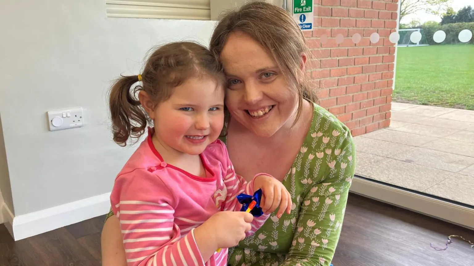 Rachel and her daughter Mia sit on foam play mats indoors. Mia is wearing a pink top and sits on Rachel’s lap holding a small toy. Behind them are a glass door, brick wall and outdoor grass.
