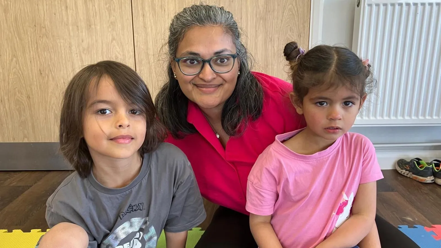 Rumayna and her two children sit on a play mat indoors. Rumayna wears a pink top, with wooden doors behind them and foam floor mats and shoes visible in the room.
