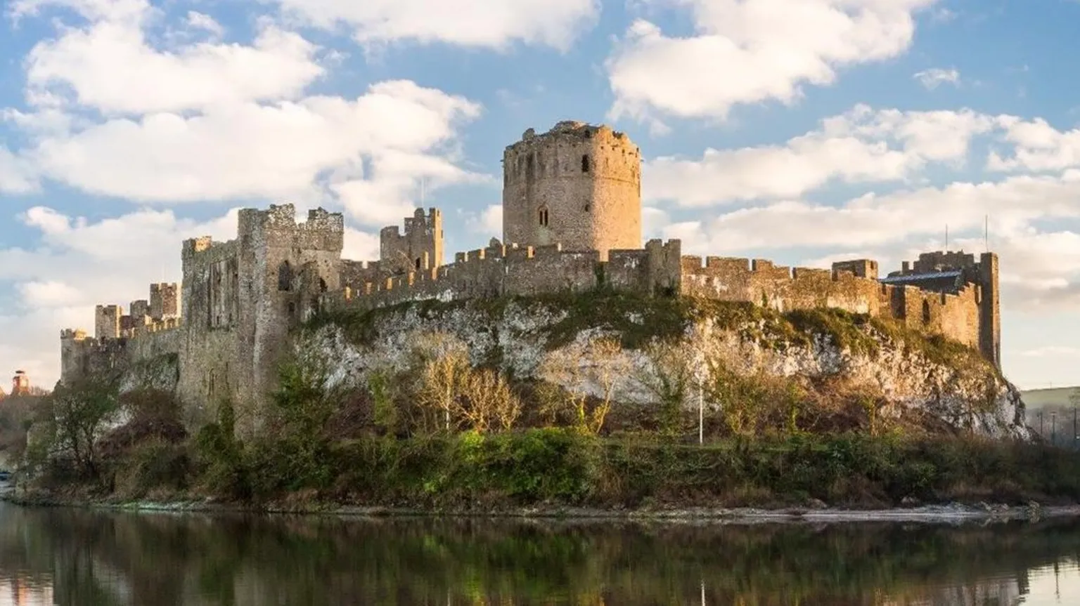 University of Aberdeen Pembroke Castle, on the banks of a river and surrounded by trees. It has tall and uneven stone walls