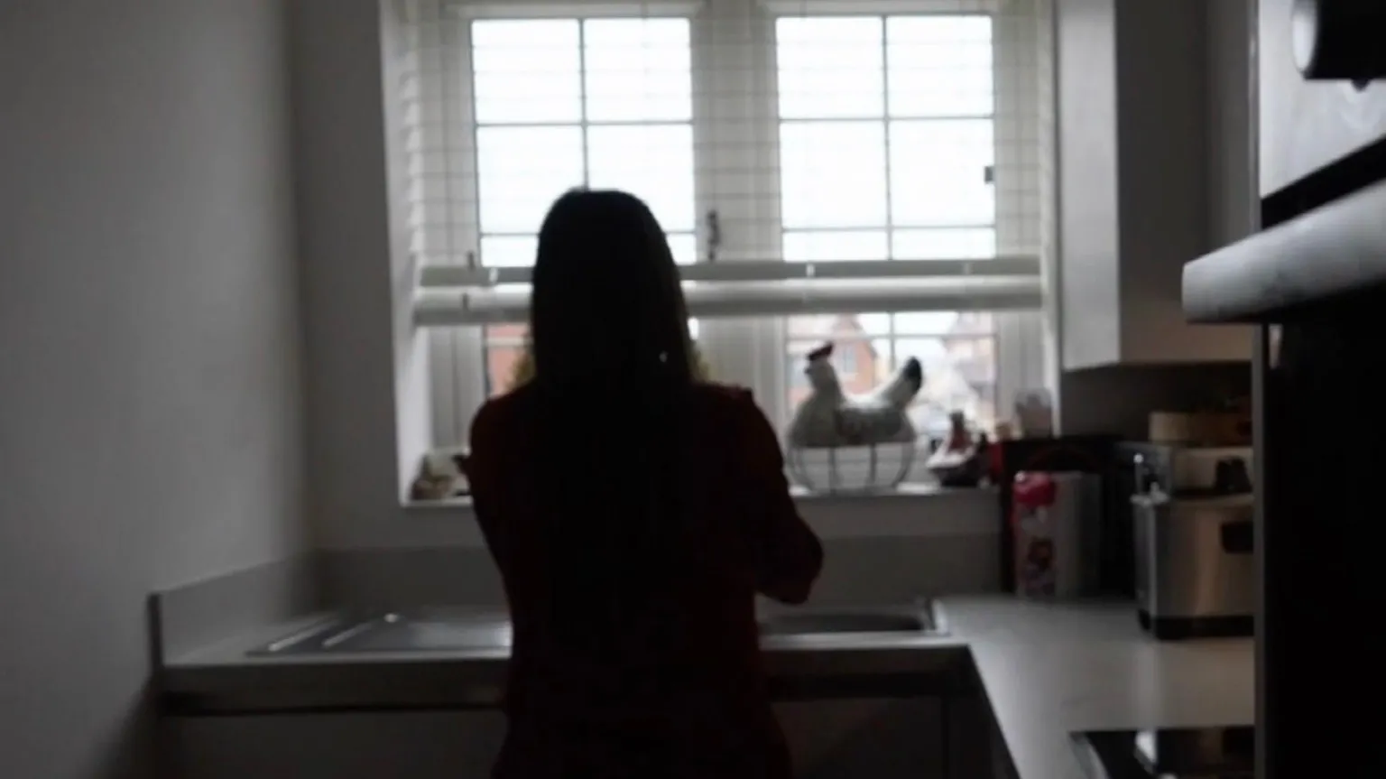 Silhouette of a young woman standing by the kitchen sink with her back to us.