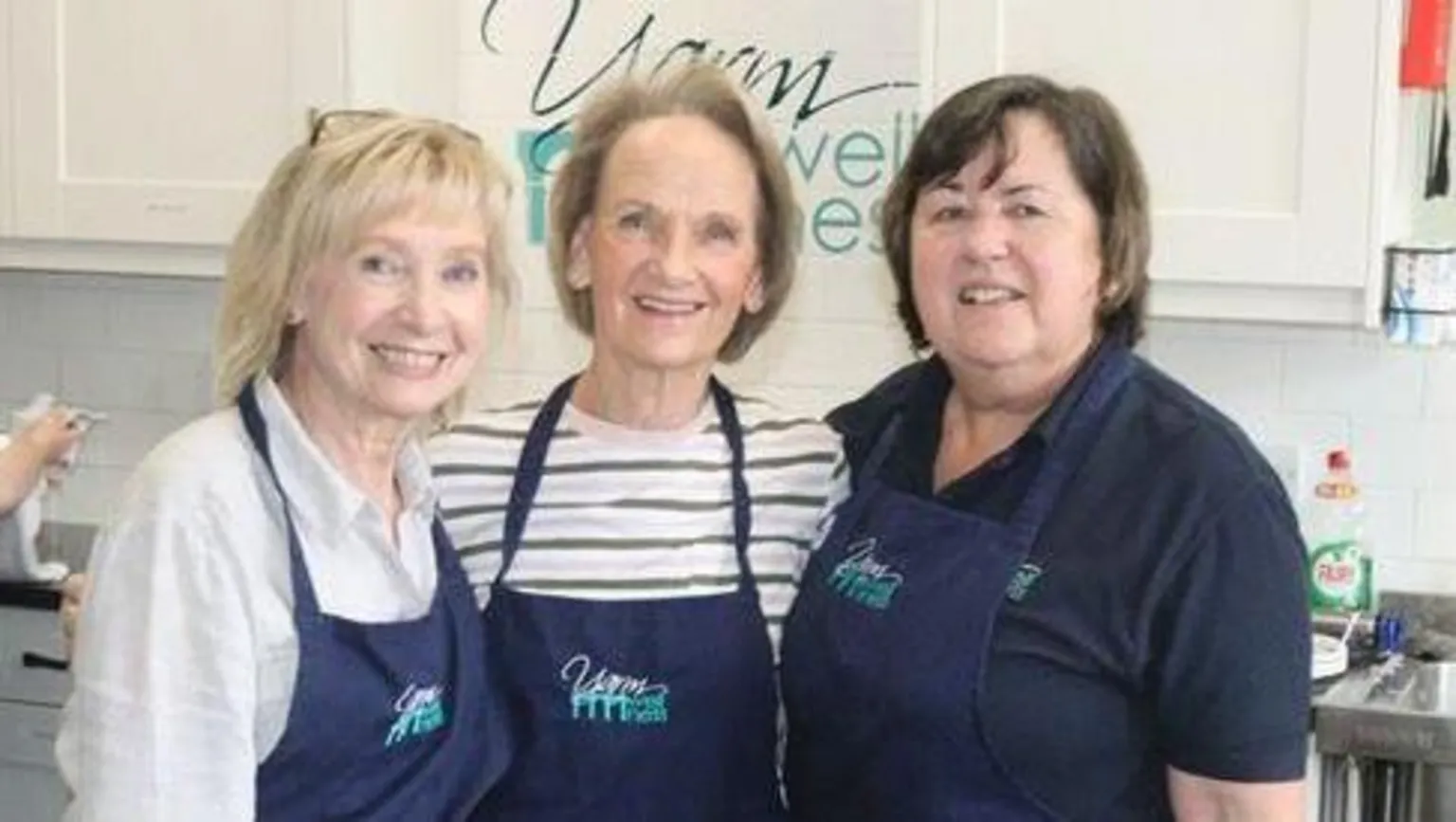 Supplied Linda Fisher is on the right with brown hair and a blue top. There are two woman next to her with blonde hair, one with a white and grey stripey top and another with a white top. They are all wearing blue aprons and standing in kitchen.
