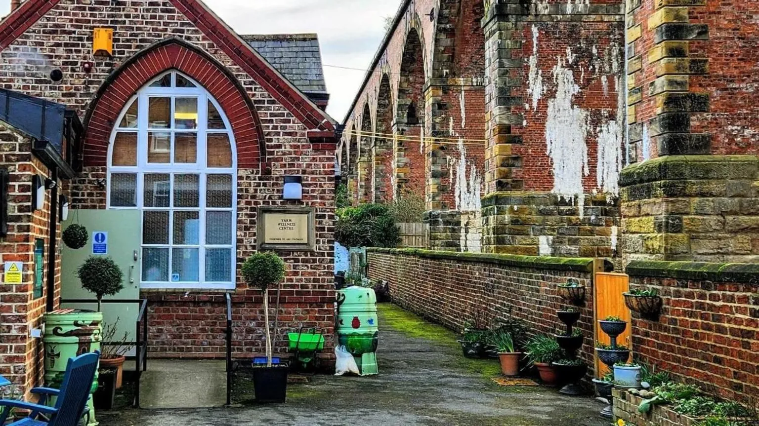 Supplied A brick building with a large white arch window. There are planters on a concrete floor. There are large stone arches to the right behind a brick wall.